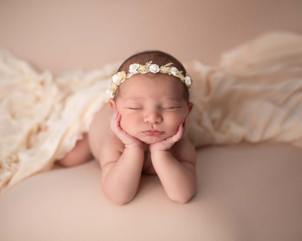 posed newborn with hands holding face on a beige blanket with headband,